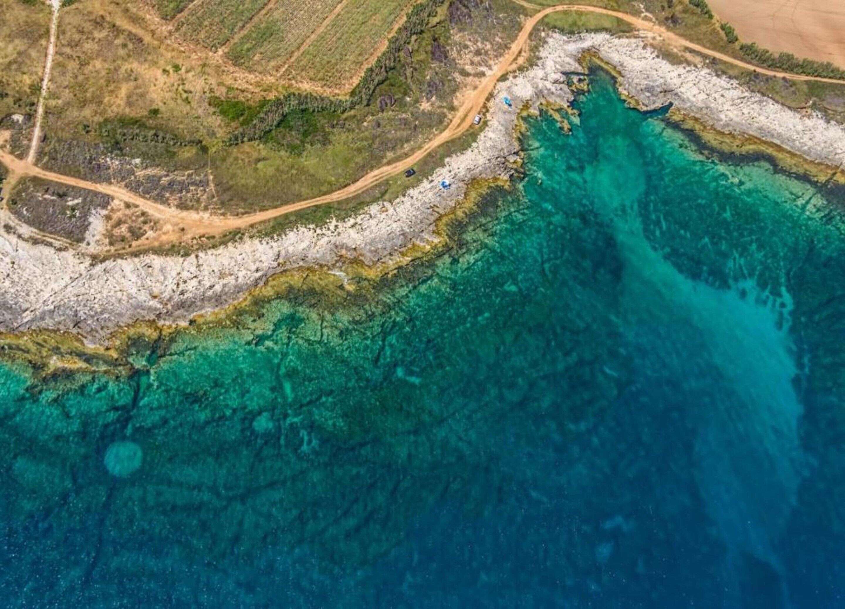 An aerial view of a coastal area with clear turquoise waters, rocky shoreline, and a dirt road.
