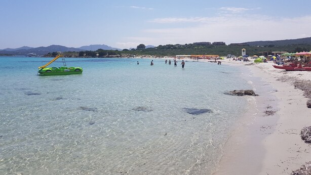 Una playa con agua turquesa cristalina, un bote inflable verde y gente disfrutando de la orilla.