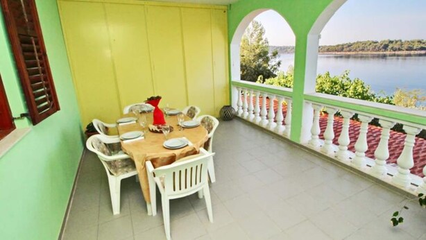 A dining area with a table set for four, surrounded by white chairs, with a view of a lake and greenery. - Apartments Bacic - Three Bedroom Apartment With Terrace and Sea View (Blato)