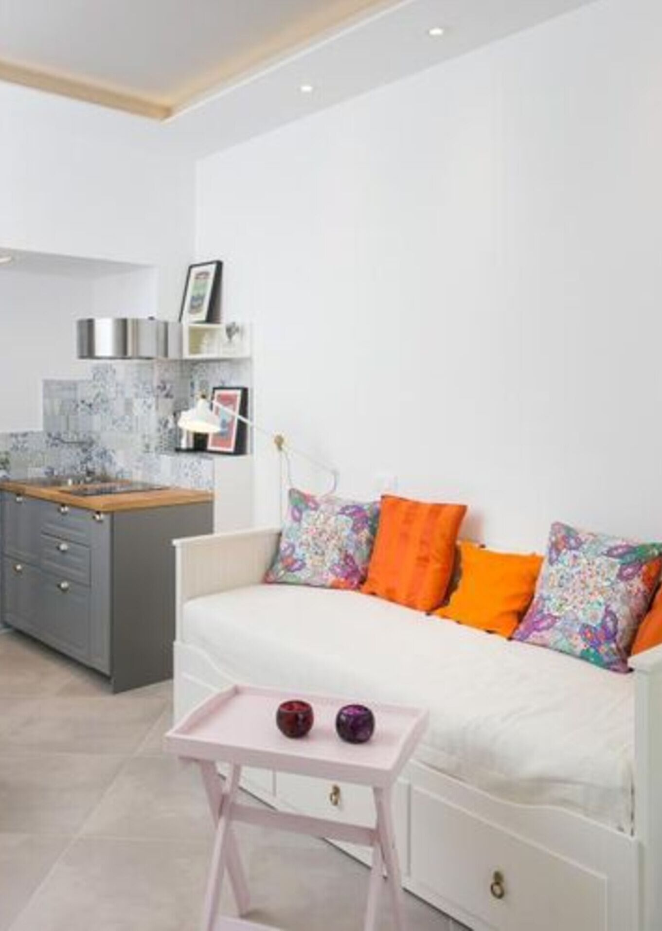 A modern kitchen with grey cabinets and a white countertop. There is a white sofa with orange and multicolored pillows, and a small pink table with two decorative items.