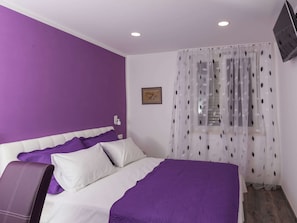 A bedroom with a purple wall, a bed with white and purple bedding, a white headboard, and a TV mounted on the wall.