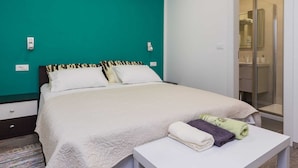 Una cama bien hecha con edredón y almohadas blancos, una mesita de noche con toallas dobladas y una pared de acento en color aguamarina.