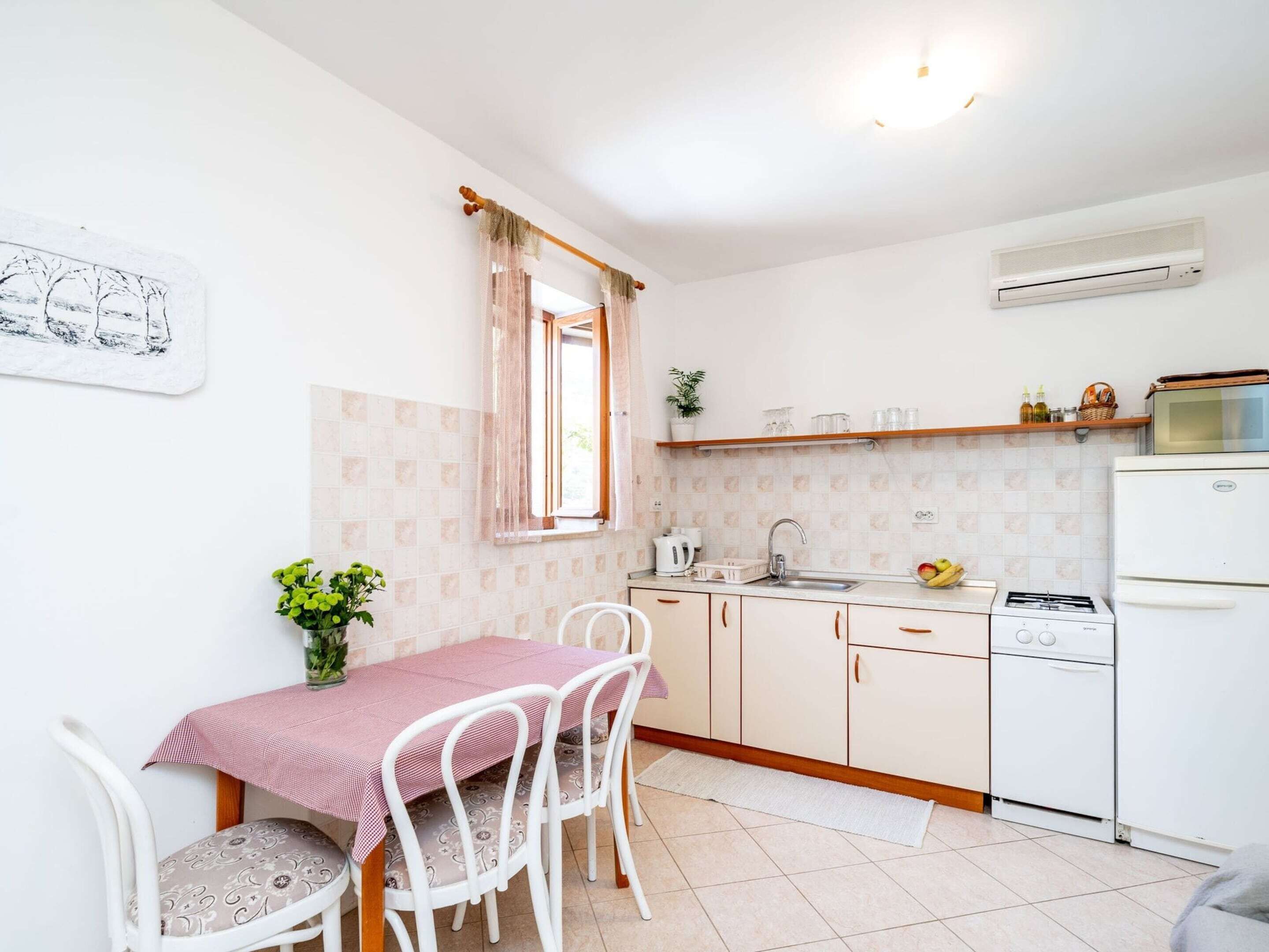 A compact kitchen with a dining area, white cabinets, a small refrigerator, and a window with sheer curtains.