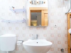 Ein Badezimmer mit einem weißen Waschbecken, einem Spiegel darüber und einer Toilette. Über dem Waschbecken befinden sich zwei transparente Kunststoffregale.