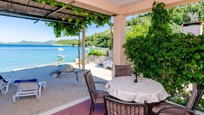 A covered outdoor seating area with a table and chairs, overlooking a beach with boats and greenery. - Apartments Kola's Mill - Comfort One Bedroom Apartment With Terrace and Sea View (Dubrovacko Primorje)