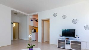 A modern living room with a flat-screen TV on a white stand, a small white table with a potted orchid, and a kitchen area in the background. - Apartments Kola's Mill - Comfort One Bedroom Apartment With Terrace and Sea View (Dubrovacko Primorje)