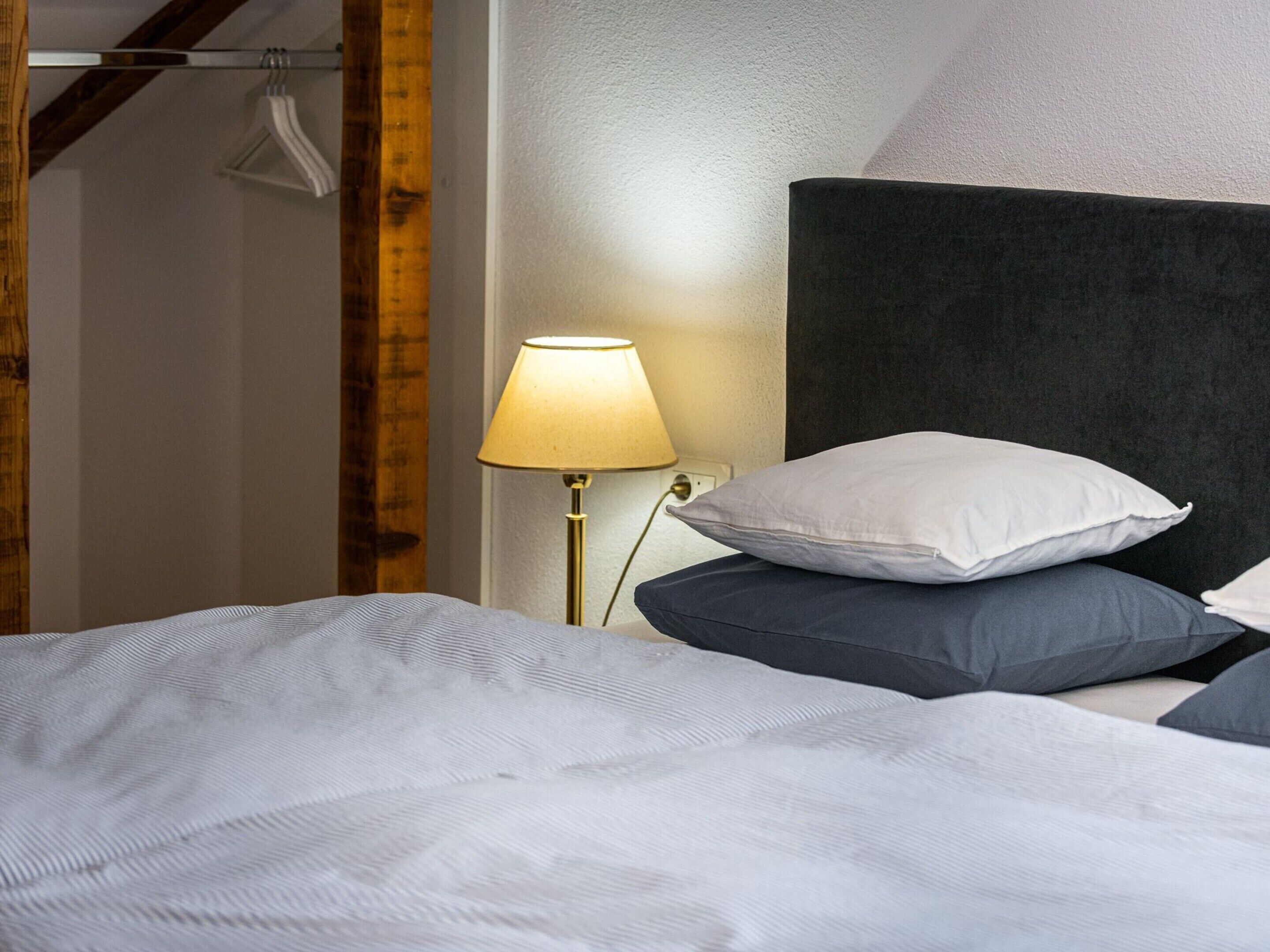 A neatly made bed with white and grey bedding, a bedside lamp, and a dark headboard.