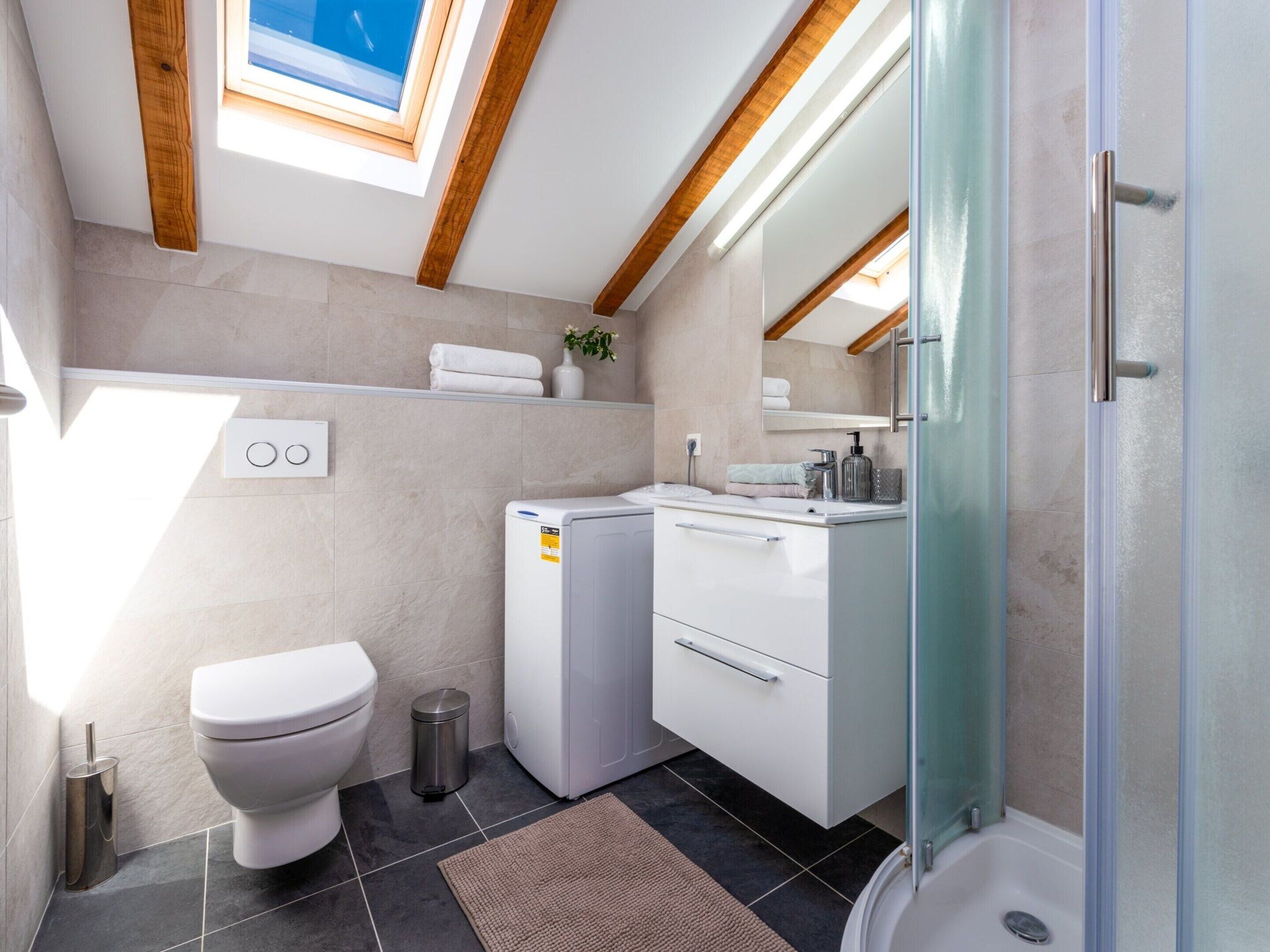 A modern bathroom with a skylight, a toilet, a washing machine, and a glass shower enclosure.