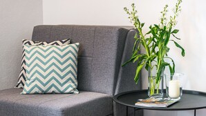 Een grijze bank met een kussen met een zigzaggeluid en een glazen vaas met groene planten en een kaars op een klein rond tafeltje.