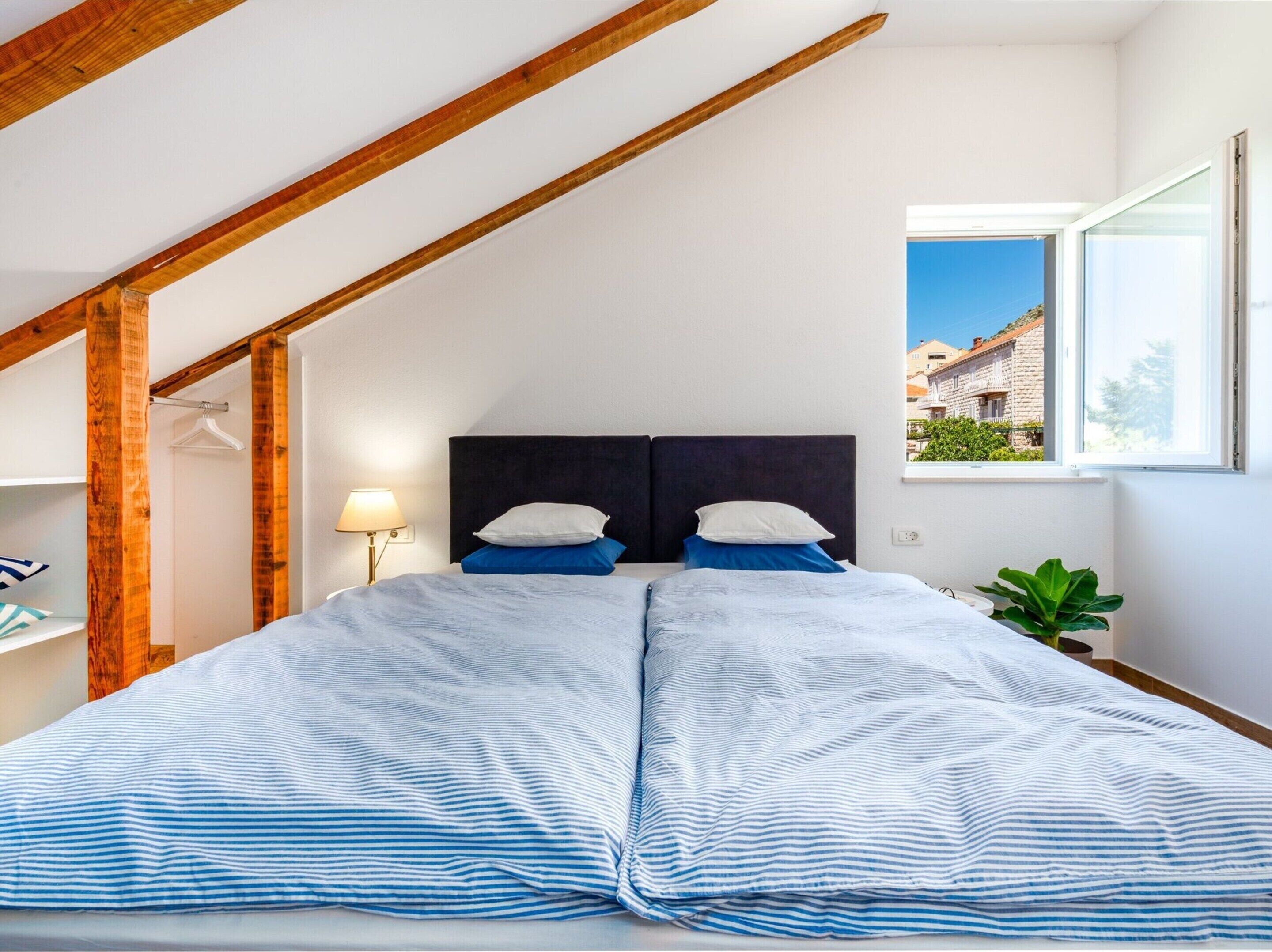 Ein Schlafzimmer mit einem großen Bett, Holzbalken, einem Nachttisch, einer Lampe und einem Fenster mit Blick auf Gebäude.