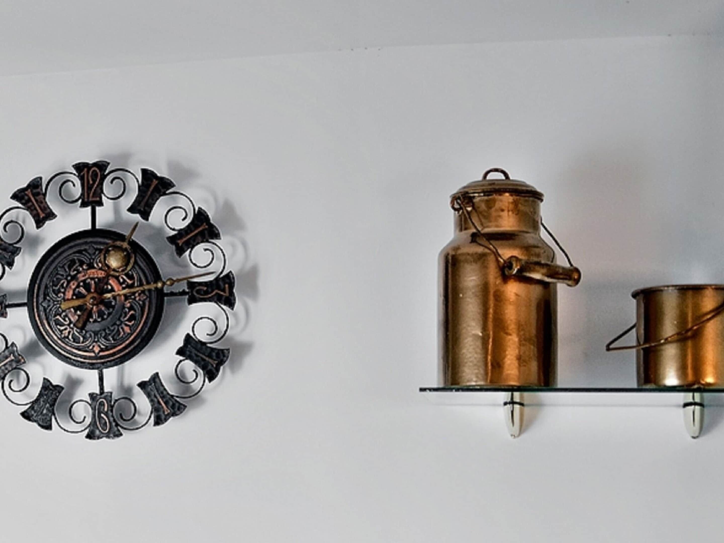 A wall-mounted clock with a decorative metal frame and a copper pot with a lid and handle on a shelf.