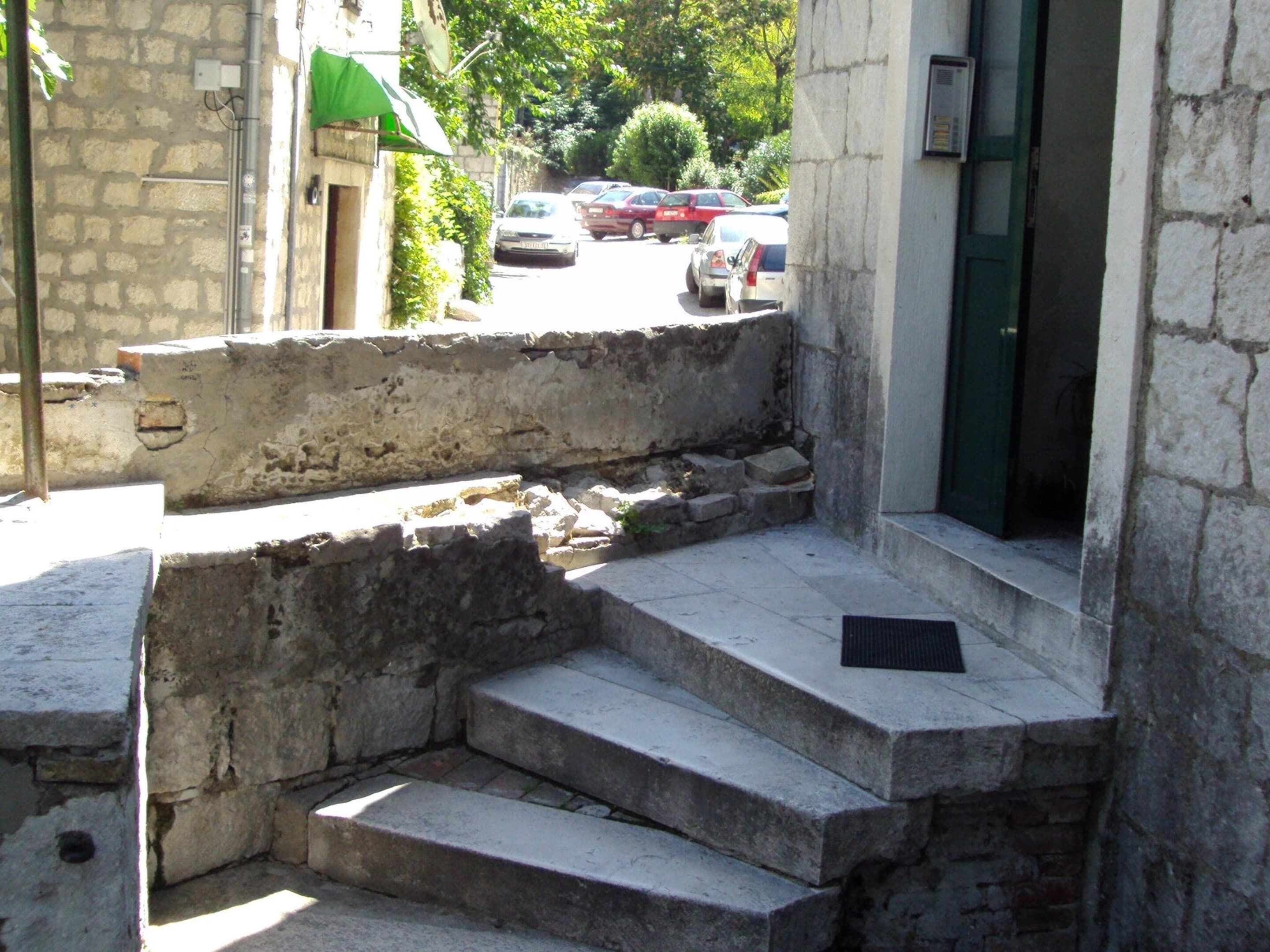 Un conjunto de escalones de piedra que conducen a la entrada de un edificio, con un toldo verde y coches aparcados al fondo.