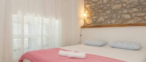 A bedroom with a stone wall, a bed with a pink blanket, and a window with sheer curtains.