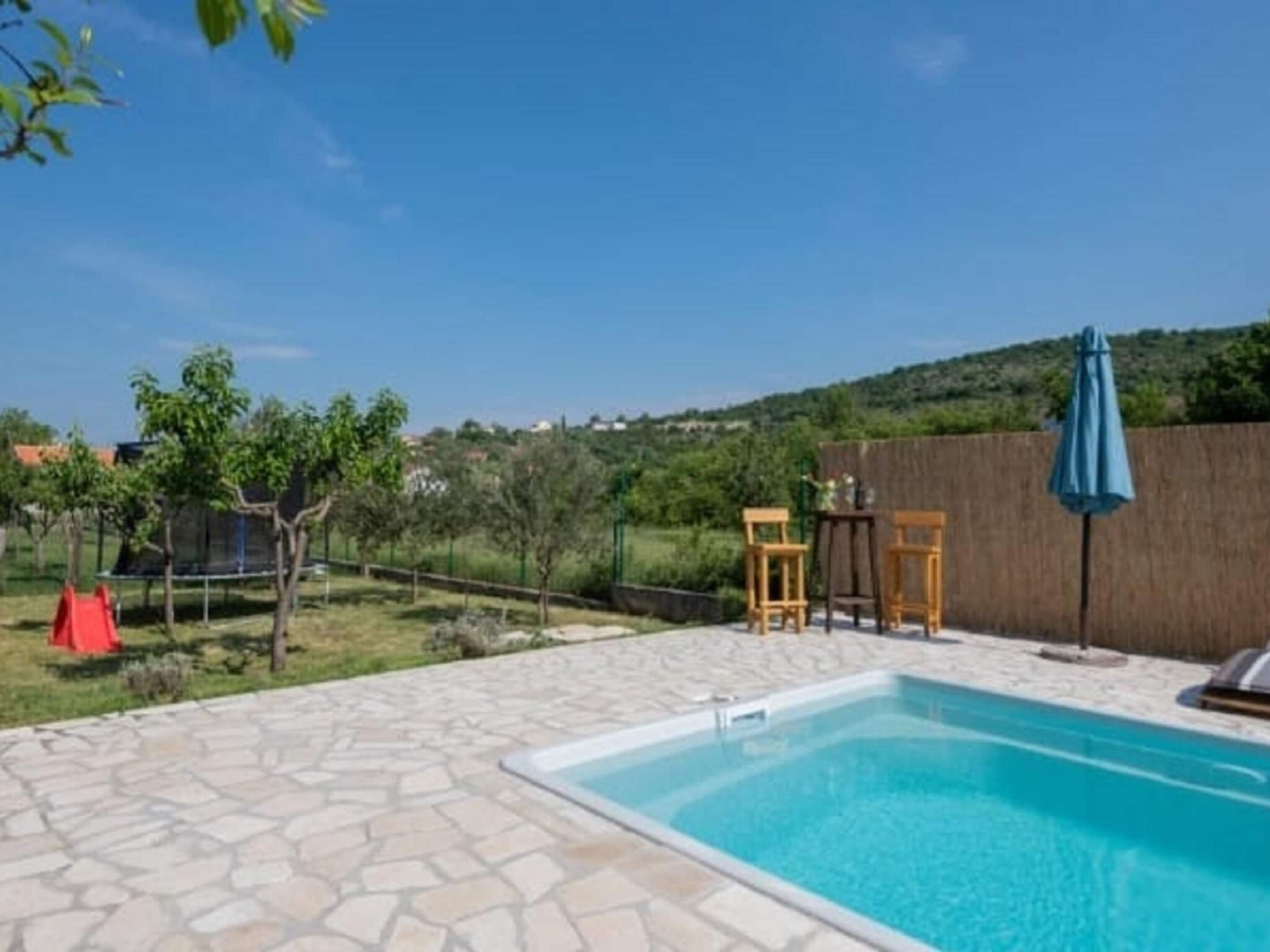 Une piscine aux bords recouverts de pierres, un toboggan rouge et deux chaises en bois sous un parasol bleu.