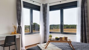 A bedroom with two large windows, a desk, a chair, and a bedside table with a tray of items.