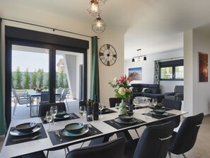 Ett modernt matområde med ett stort bord dukat för sex personer, en soffa i bakgrunden och utsikt över utomhusmiljön genom en glasdörr.