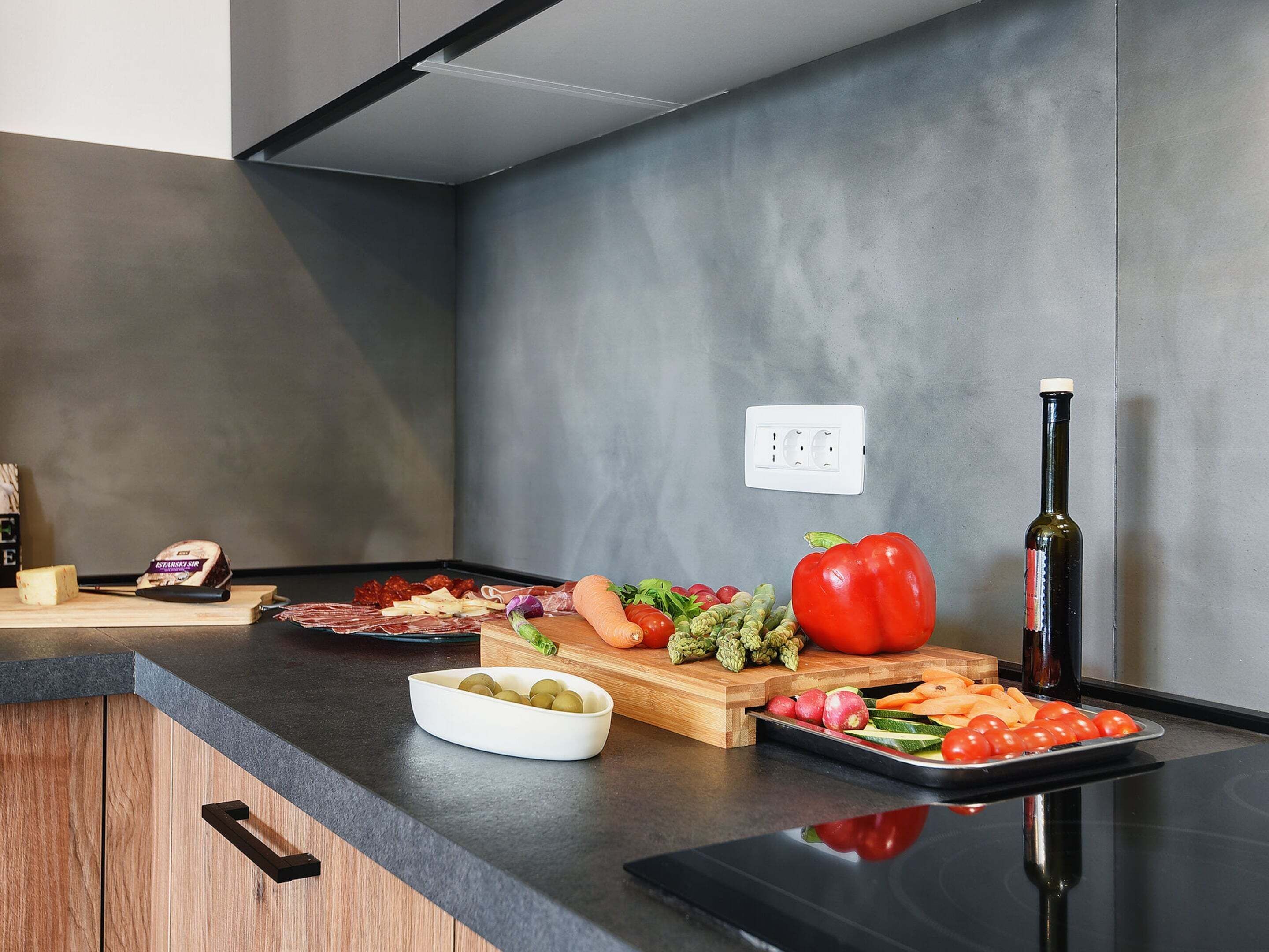 Eine moderne Küche mit grauer Wand, einer Holz Arbeitsplatte und verschiedenen Lebensmitteln, darunter eine rote Paprika, Oliven und eine Flasche Olivenöl.