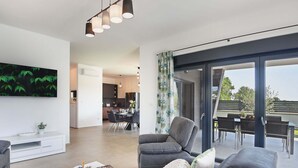 A modern living room with a grey sofa, a coffee table, and a TV. There is a dining area with a table and chairs, and a kitchen in the background.