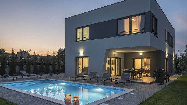 A modern two-story house with a swimming pool, outdoor seating, and a well-lit patio area.