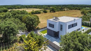 A modern two-story house with a flat roof, surrounded by lush greenery and a large open field.