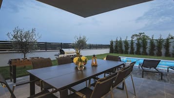 An outdoor patio with a table and chairs, a grill, a pool, and a view of the ocean.