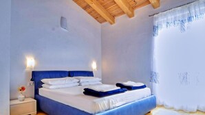 Une chambre avec un plafond en bois, un lit avec une literie bleue, une table de chevet avec un vase et une fenêtre ornée de rideaux légers.