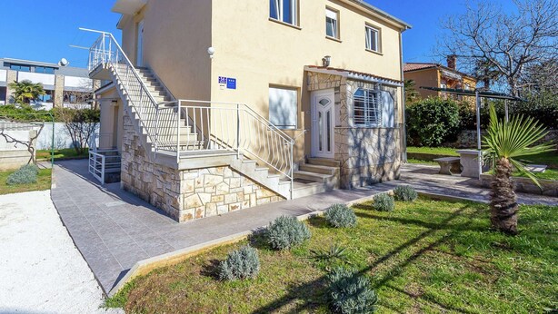 Un bâtiment à deux étages, avec un escalier en pierre menant à l’entrée, un petit porche et un ciel bleu clair en arrière-plan.