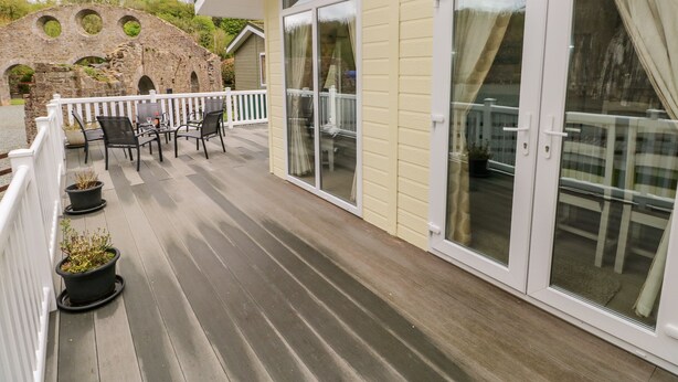 A covered outdoor area with a wooden deck, a white railing, and a seating area with chairs and tables. - Seacroft (Narberth)