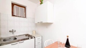 A small kitchen with white cabinets, a sink, and a window with a curtain. There is a table with a tablecloth and a bottle of wine. - Apartment Dalmatin Zuljana - Two-bedroom Apartment 2 (Ston)