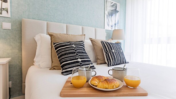 Una cama impecablemente arreglada con una bandeja de desayuno con cruasanes, zumo de naranja y café, frente a una pared azul claro y un cabecero moderno.