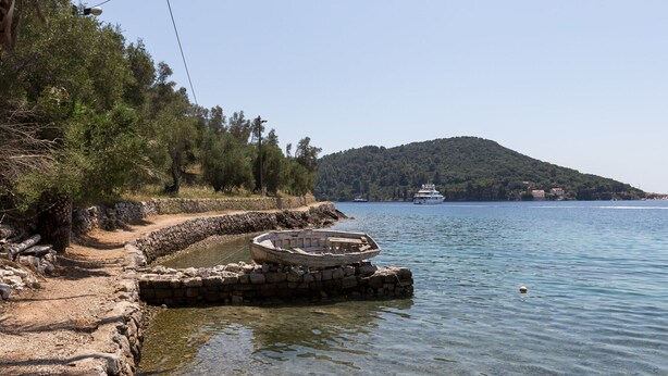 A coastal scene with a stone pathway leading to a dock, a small boat, and a yacht in the background.