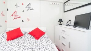 Une chambre à coucher avec un lit, deux oreillers rouges, une commode blanche surmontée d’un téléviseur à écran plat, et un mur décoré d’autocollants représentant des papillons.
