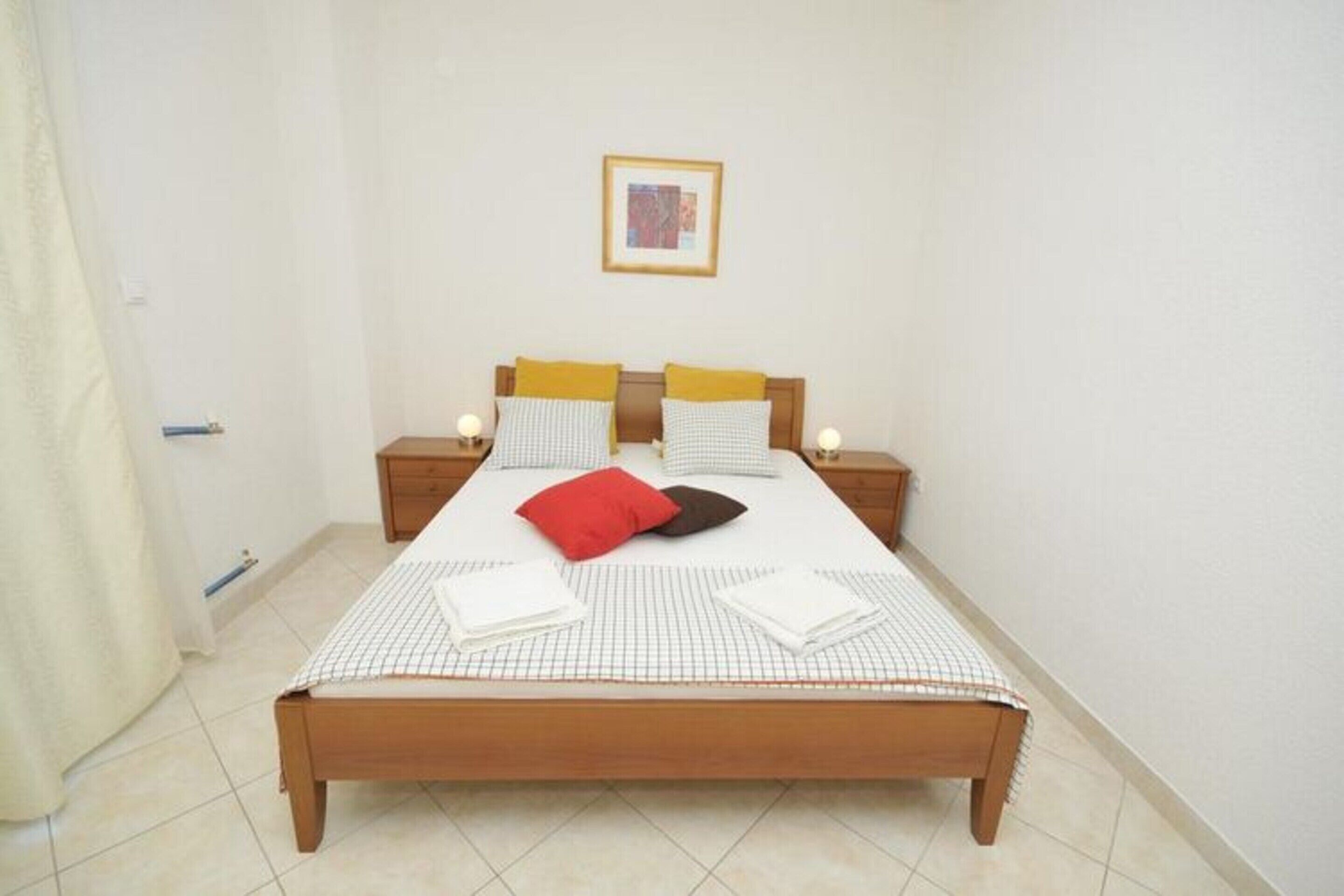 A single bed with a checkered bedspread, two bedside tables with lamps, a framed picture on the wall, and a curtain on the left.