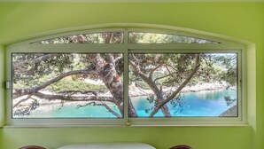A room with a large window overlooking a turquoise body of water, surrounded by trees. - Seafront Apartments Olivera-standard One Bedroom Apartment With Balcony and Sea View (Blato)