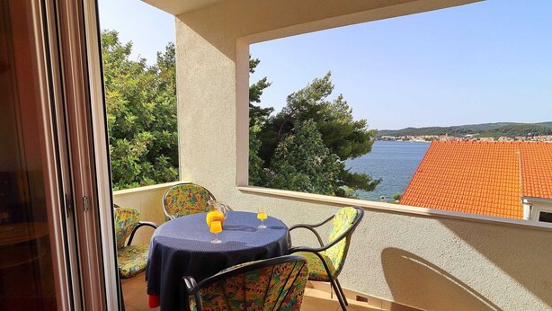 A balcony with a table and chairs, a view of the sea, and a tiled roof in the background.
