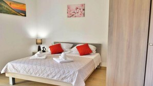 A neatly made bed with white and red pillows, a bedside lamp, and a painting of a sunset on the wall. - Fancy Apartments Dream - Modern One Bedroom Apartment With Balcony and Sea View (Smokvica)