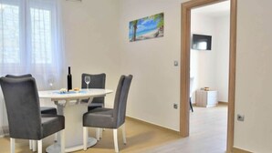 A dining area with a round table, four grey upholstered chairs, a wine bottle, and a glass. A painting of a beach scene hangs on the wall. - Fancy Apartments Dream - Modern One Bedroom Apartment With Balcony and Sea View (Smokvica)