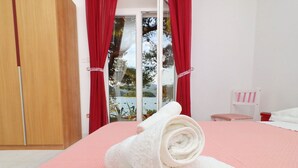 A bedroom with a pink bedspread, a white rolled towel, a wooden wardrobe, and a window with red curtains. - Apartments More & Ja - Superior One Bedroom Apartment With Terrace and Sea View (Ston)