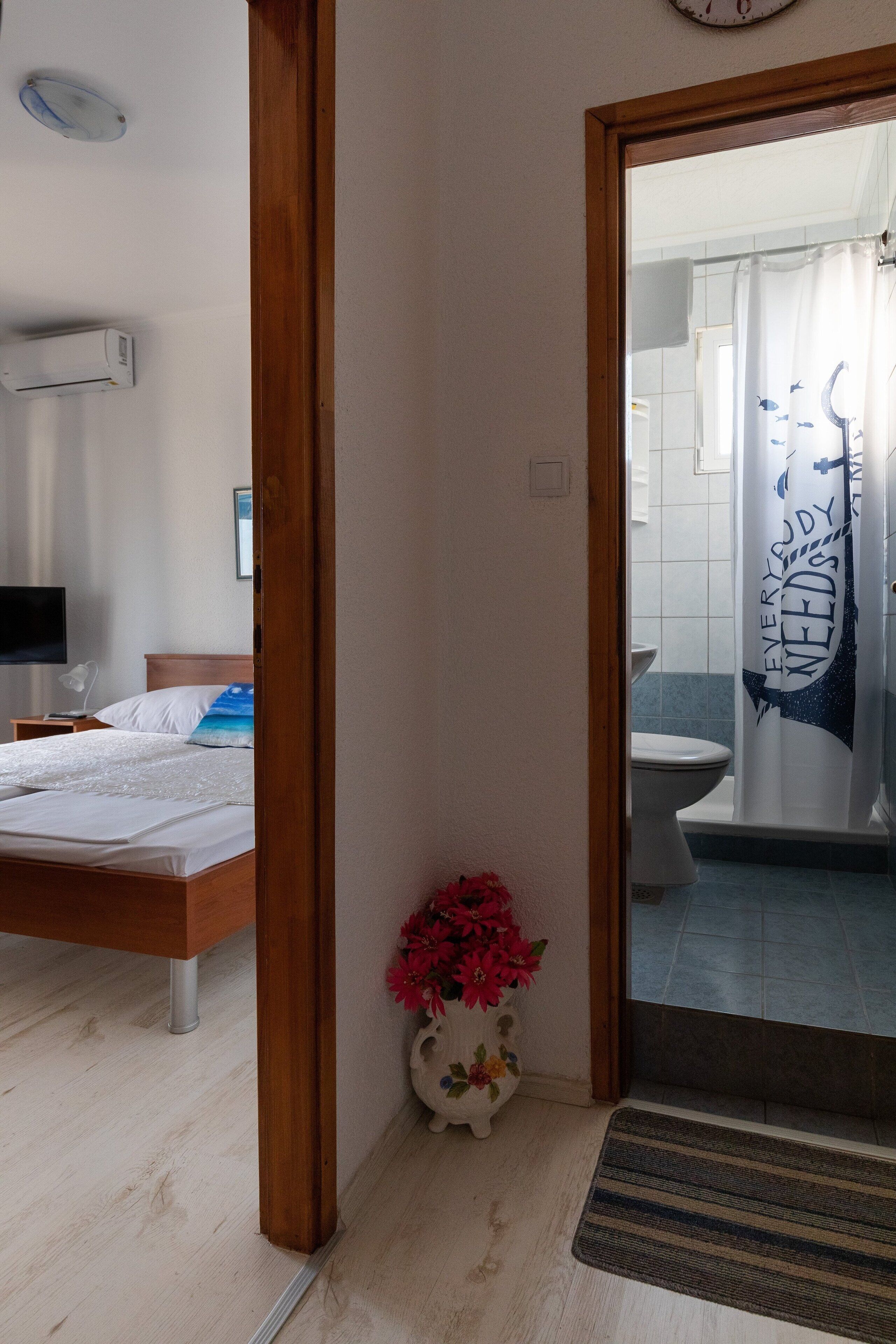 A hotel room with a wooden bed, a bedside table with a lamp, a small vase with flowers, and a view into the bathroom through a glass door.