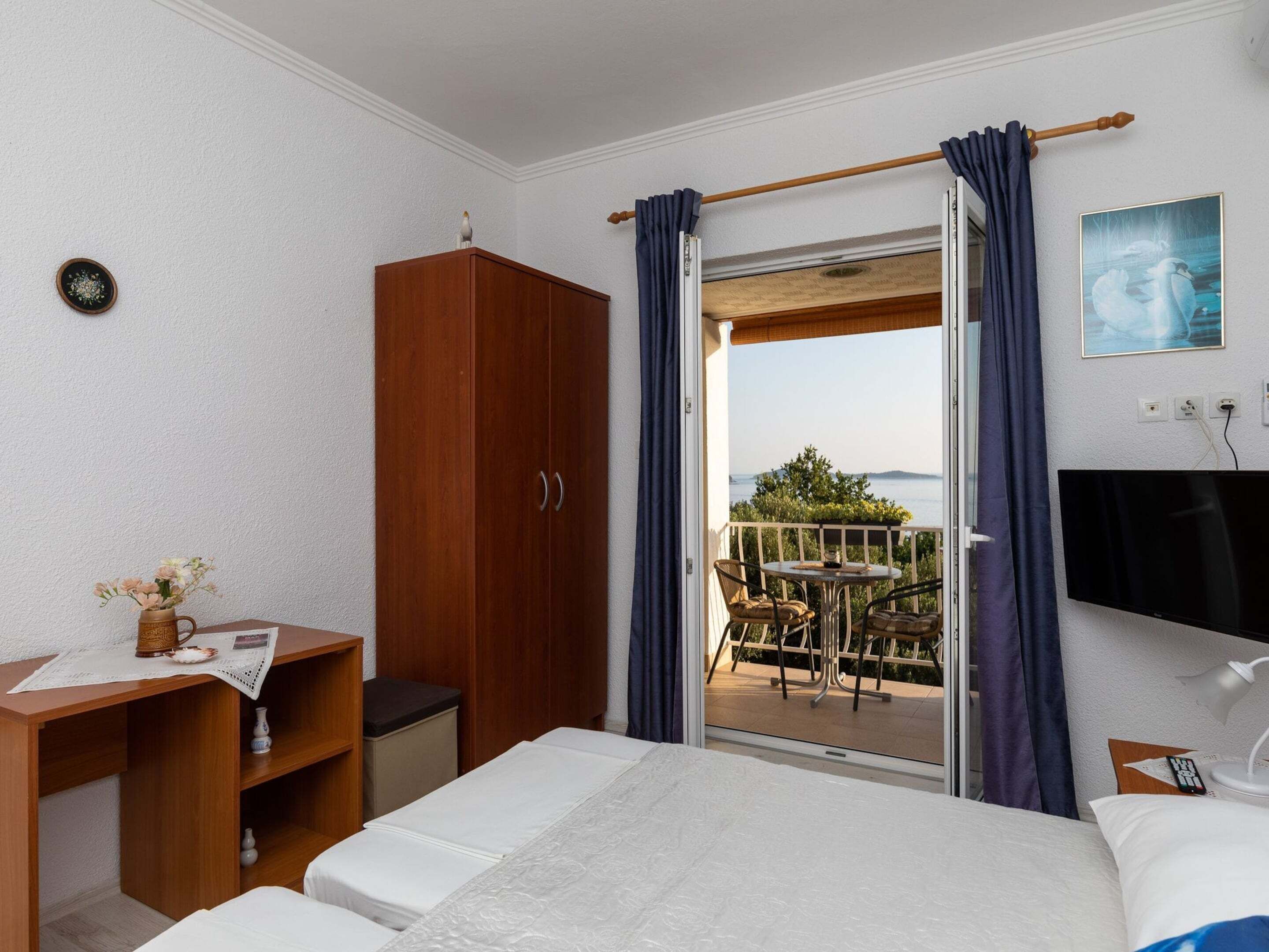 Quarto de hotel com cama, escrivaninha, guarda-roupa, varanda com mesa e cadeiras, e televisão na parede.