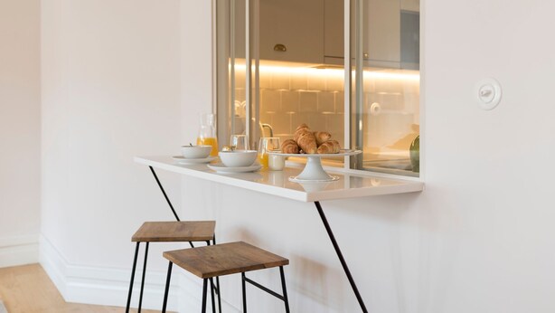 Een moderne keuken met een wit aanrecht, twee houten krukken en ontbijtartikelen zoals croissants en sinaasappelsap.