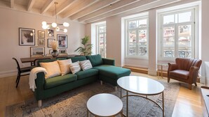 Een woonkamer met een groene hoekbank, een ronde salontafel en een eetgedeelte met een tafel en stoelen. Grote ramen bieden uitzicht op de stad.