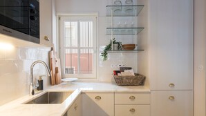 Een moderne keuken met een ingebouwde kookplaat, een spoelbak en een raam. Boven het aanrecht zijn glazen planken en een mand met spullen staat er.