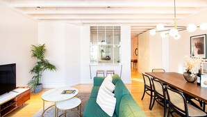 Een moderne woonkamer met een groene bank, een klein rond tafeltje, een televisie en een eethoek met een houten tafel en stoelen.