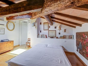 A bedroom with a wooden ceiling, a bed, a dresser, a chair, and a wall-mounted shelf with books and decorative items. - Pisa Getaway by the Tower (Pisa)