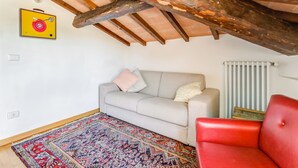 Une pièce avec un plafond haut, des poutres apparentes, un canapé gris, un fauteuil rouge, un tapis à motifs et un radiateur.