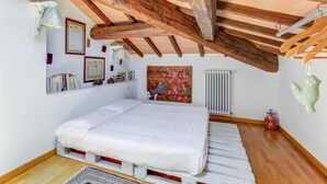 Une petite pièce agréable, avec un plafond en bois, un lit, un radiateur et une étagère fixée au mur, où sont posés des livres et des objets décoratifs.