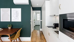 Una cocina moderna con armarios blancos, una encimera negra y un horno empotrado. Hay un comedor con una mesa y sillas de madera, y un pasillo que conduce a un baño.