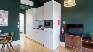 A compact kitchen with white cabinets, a microwave, and a small coffee maker. There's a wooden dining table with chairs, a television on a wooden stand, and a fire extinguisher on the wall. - Lóios 66, 4º by LovelyStay (Porto)