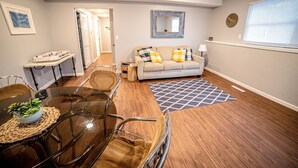 A modern living room with a glass dining table, a sofa with patterned cushions, and a wooden floor.